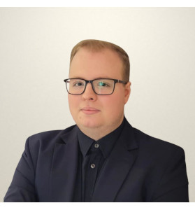 A young man with light, short hair. Wearing black-rimmed glasses. Dressed in a black shirt buttoned up to the neck and a black jacket. A light background behind. The man is looking straight into the camera lens.