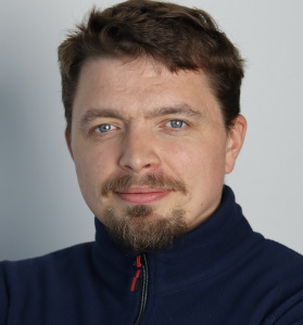 Male. Only the bust is visible. Dark hair. Moustache and beard. A gentle smile. Dressed in a navy blue sweatshirt with the zipper pulled up to the neck.