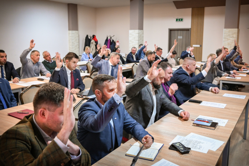 The photo shows a group of people sitting at tables in a conference or meeting room, attending a meeting of the Association of Polish Cities. Everyone is raising their hands as they take part in the vote. The individuals are dressed formally, in suits and jackets. The tables are arranged in rows and there are various items such as documents and electronic devices.